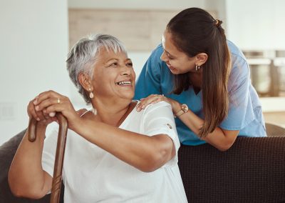 Nurse leaning down to speak with a resident seated on a couch holding a cane