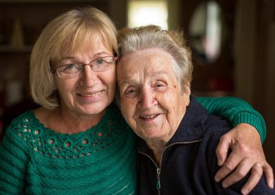 Daughter wearing glasses and a green sweater with her arm around her mom wearing a black track suit